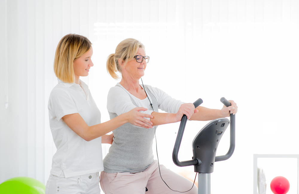 Ältere Frau trainiert auf Ergometer mit Unterstützung einer Therapeutin