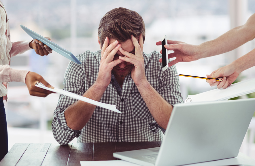 Gestresster Mann am Laptop hält sich den Kopf während Kollegen ihn ansprechen