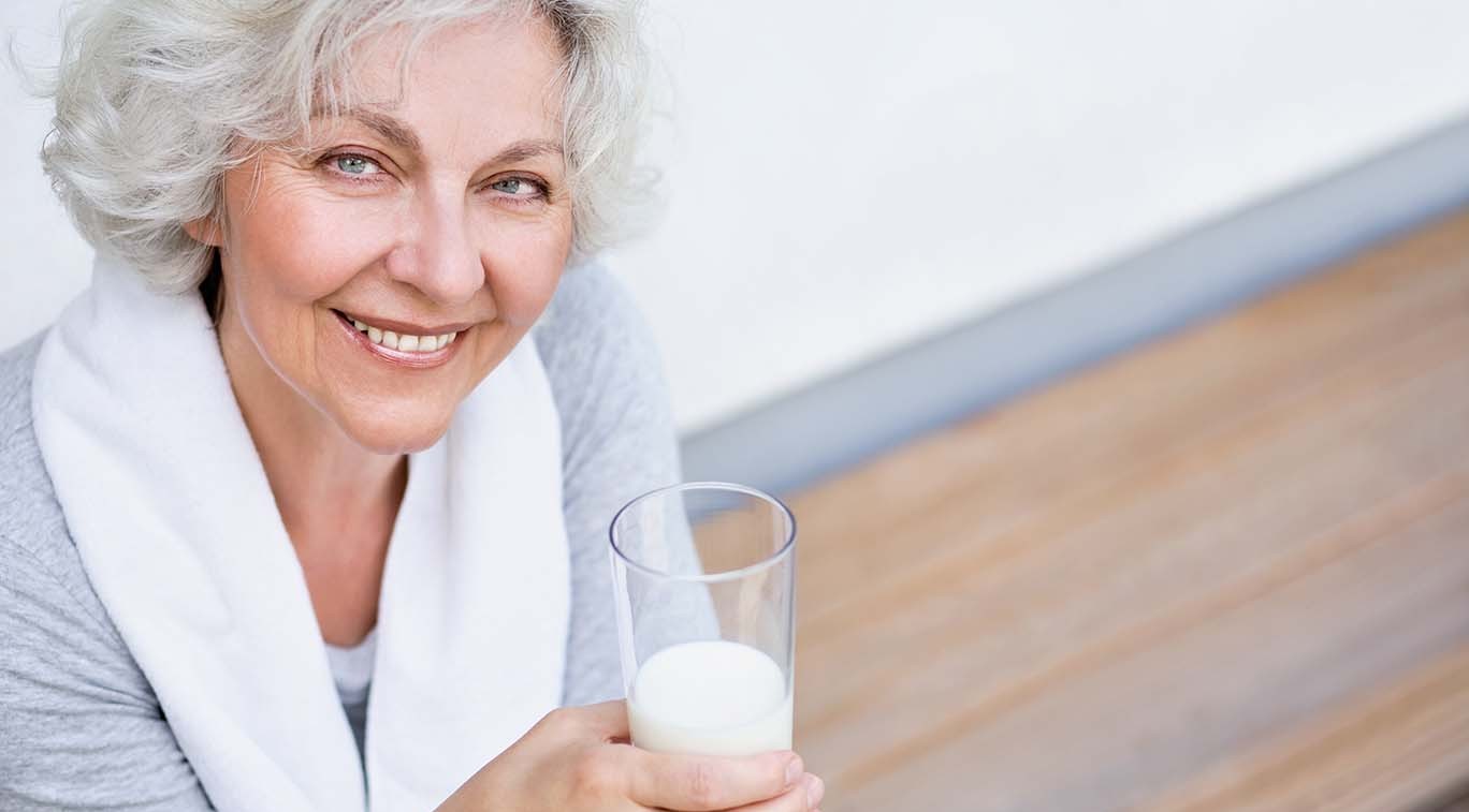 Ältere Frau mit grauen Haaren hält ein Glas Milch und lächelt. Eine kalziumreiche Ernährung, z. B. durch Milchprodukte, hilft zur Vorbeugung von Osteoporose und stärkt die Knochengesundheit.