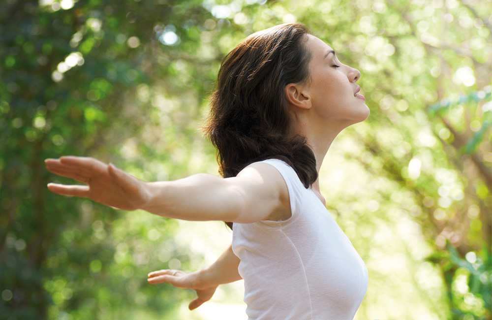 Frau steht im Grünen mit ausgebreiteten Armen und atmet bewusst ein