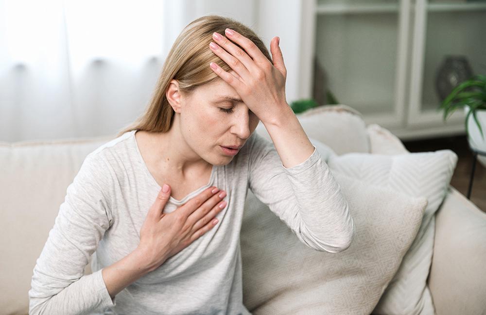 Frau sitzt erschöpft auf dem Sofa und hält sich an Brust und Stirn