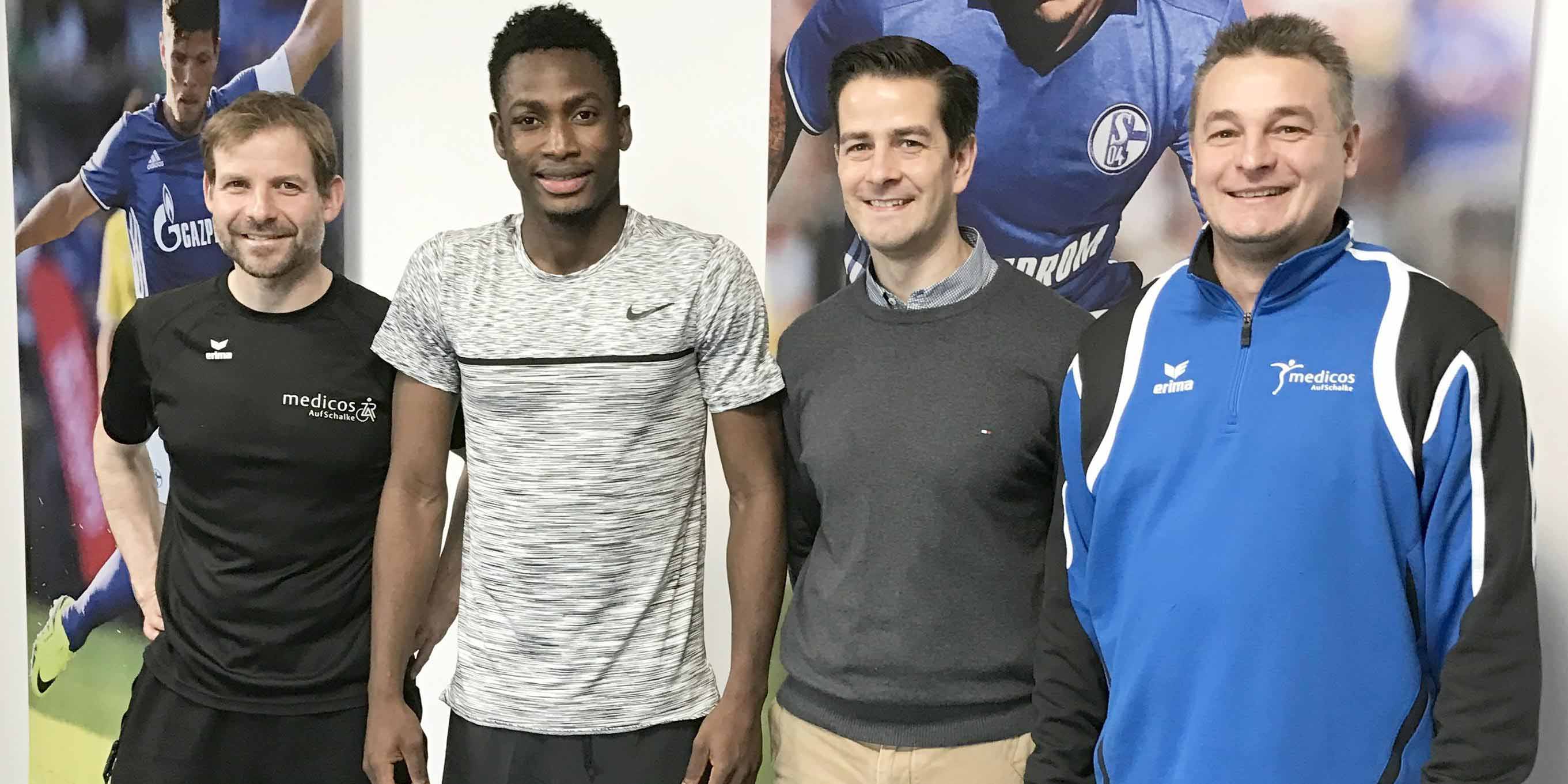 Ein Gruppenfoto mit einem Physiotherapeuten-Team, darunter ein Athlet in Sportkleidung, vor einer Wand mit Schalke 04-Motiven.