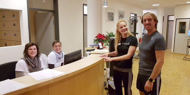 Ein Empfangsbereich mit zwei Mitarbeiterinnen hinter einem Tresen und zwei Trainierenden in Sportkleidung, die lächelnd auf den Tresen zeigen.