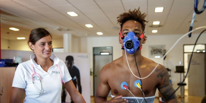 Ein Sportler mit Atemmaske und Brustelektroden während eines Leistungstests, begleitet von einer medizinischen Fachkraft mit Stethoskop.
