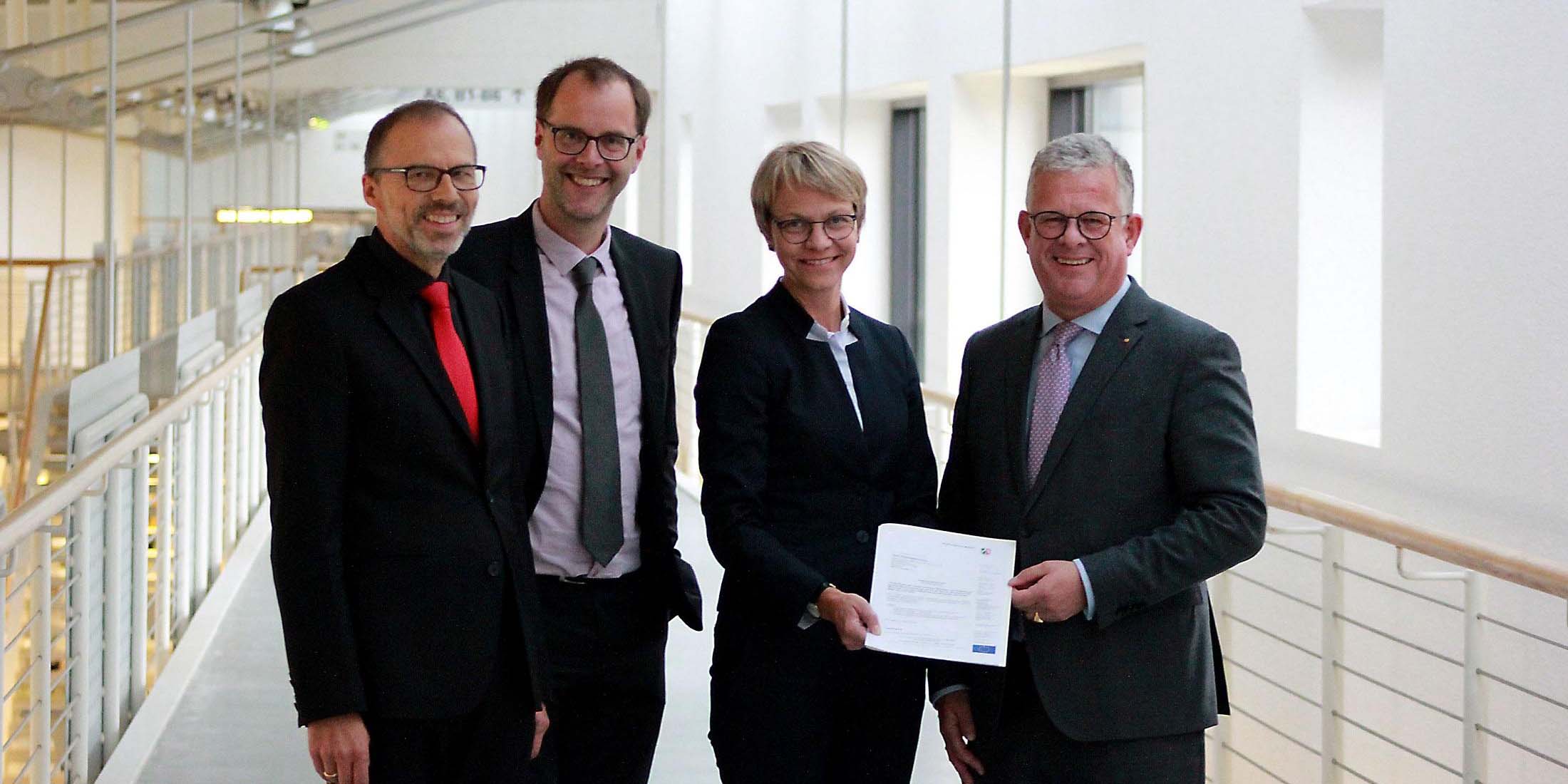 Vier Personen in Businesskleidung posieren mit einem Dokument in einem modernen Gebäude.